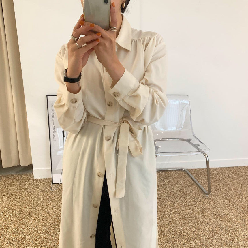 Woman in a cream-colored button-down dress with a belt, holding a phone and taking a selfie.