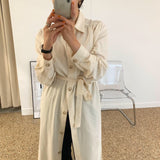 Woman in a cream-colored button-down dress with a belt, holding a phone and taking a selfie.