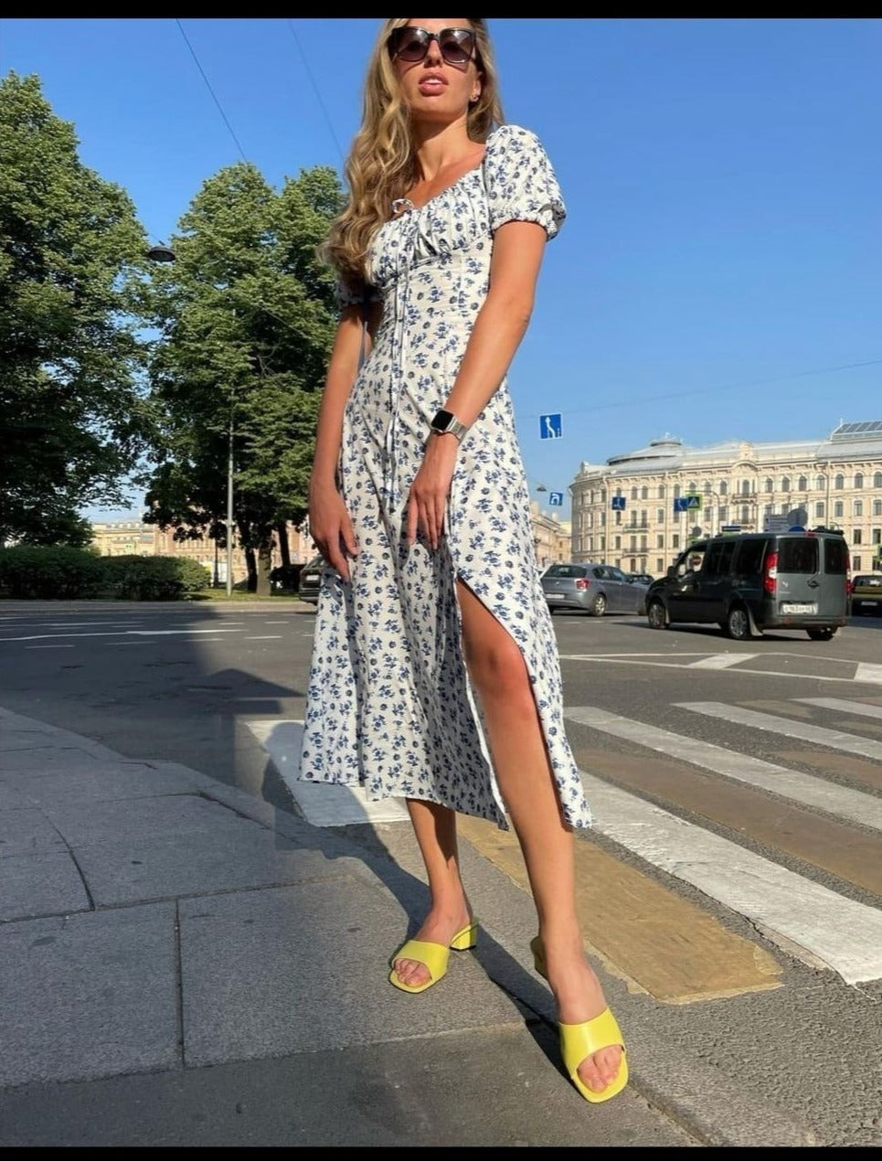 A woman in a white and blue floral print midi dress with a high slit poses on a city street with a crosswalk.