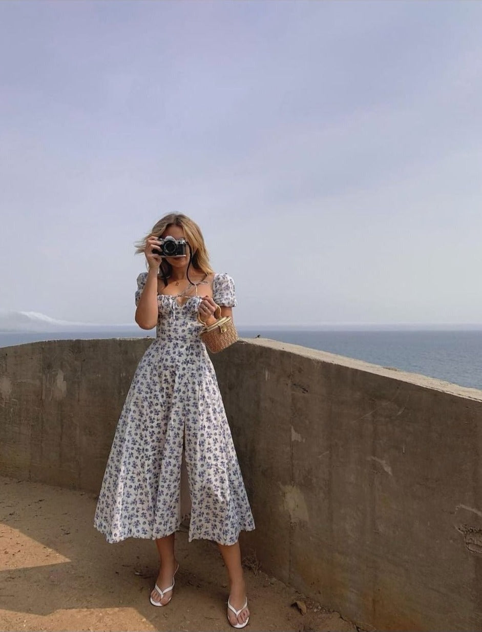 A woman in a white and blue floral print midi dress with a high slit takes a photo with a vintage camera by the ocean.