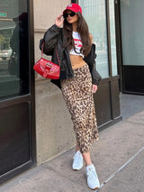 A woman wearing a white crop top, black bomber jacket, and a long leopard print skirt, with a red purse and red baseball cap.