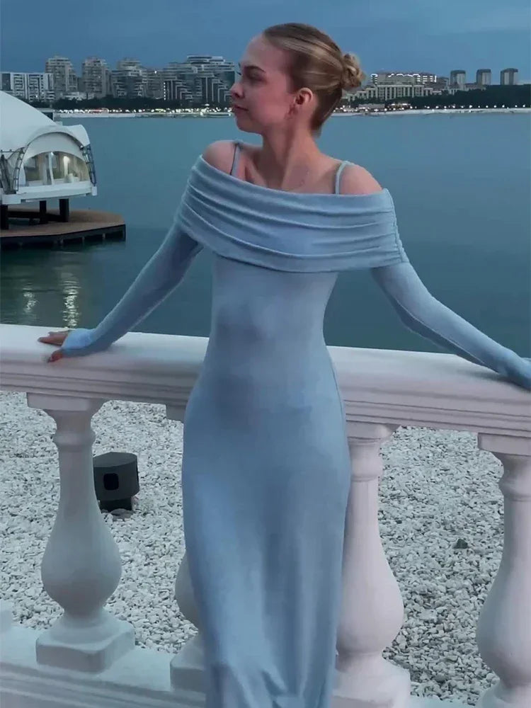 A young woman wearing a light blue off-the-shoulder dress with long sleeves, leaning on a white railing with a cityscape and ocean in the background.