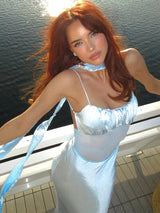 Close-up of a woman with auburn hair in a light blue satin slip dress with a ruched bust and sheer midsection, with the sun glinting off the water.