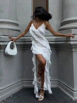 A woman in a white ruffled maxi dress leans against a stone wall, holding a white handbag.