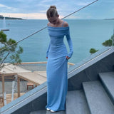 A young woman wearing a long, light blue off-the-shoulder dress with long sleeves, standing on outdoor stairs with the ocean in the background.