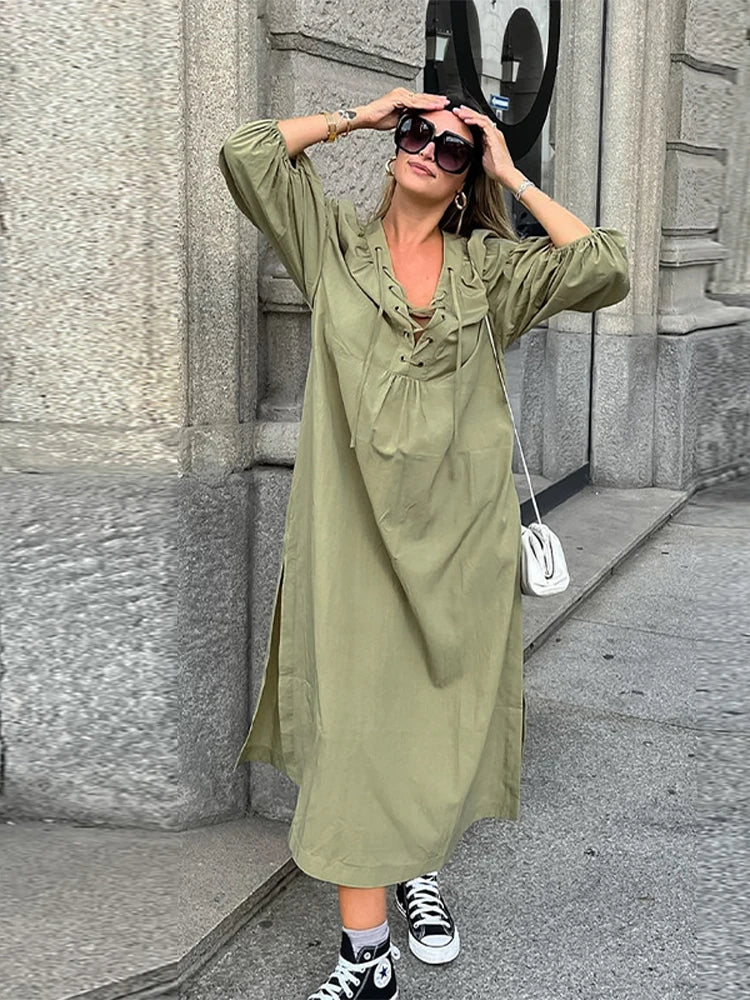 Woman in an olive green maxi dress with puff sleeves and a lace-up neckline, posing with hands on sunglasses, wearing black high-top sneakers.