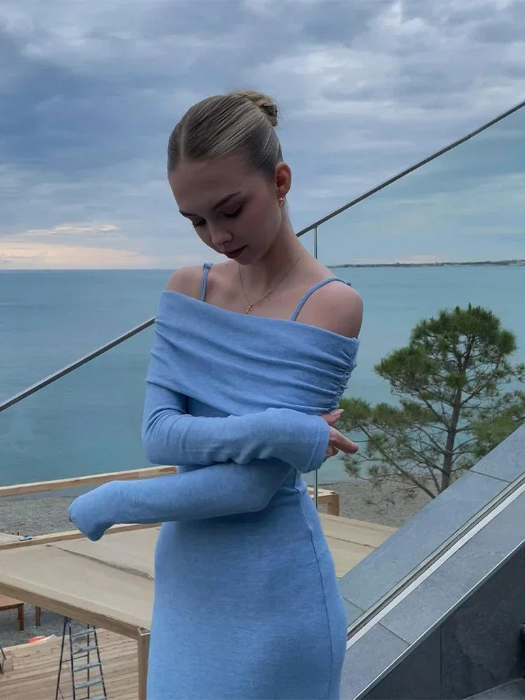 A close-up of a young woman wearing a light blue off-the-shoulder dress with ruching detail, looking down, with the ocean in the background.