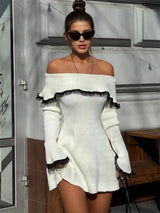 A woman in sunglasses wearing a white off-the-shoulder ribbed knit dress with black lace trim, sitting on a bench.