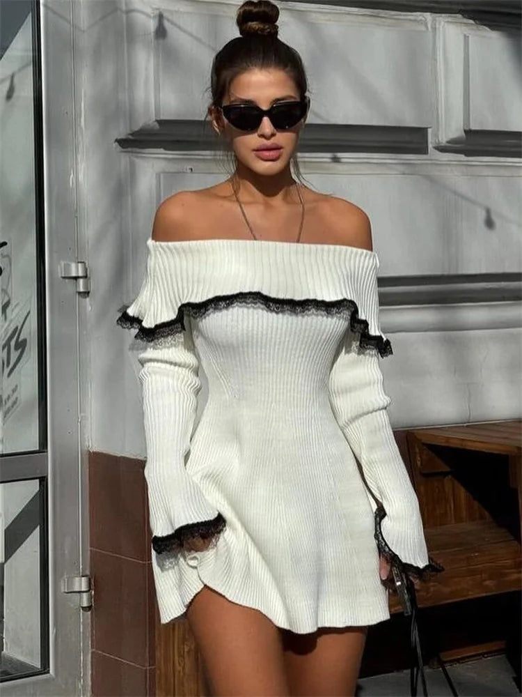 A woman in sunglasses wearing a white off-the-shoulder ribbed knit dress with black lace trim, sitting on a bench.