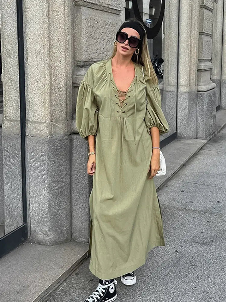 Woman wearing a long olive green dress with puff sleeves and a lace-up neckline, paired with black high-top sneakers and a white crossbody bag.