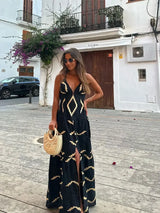 Woman in a black maxi dress with a gold geometric print, holding a straw bag and standing on a street with a building in the background.