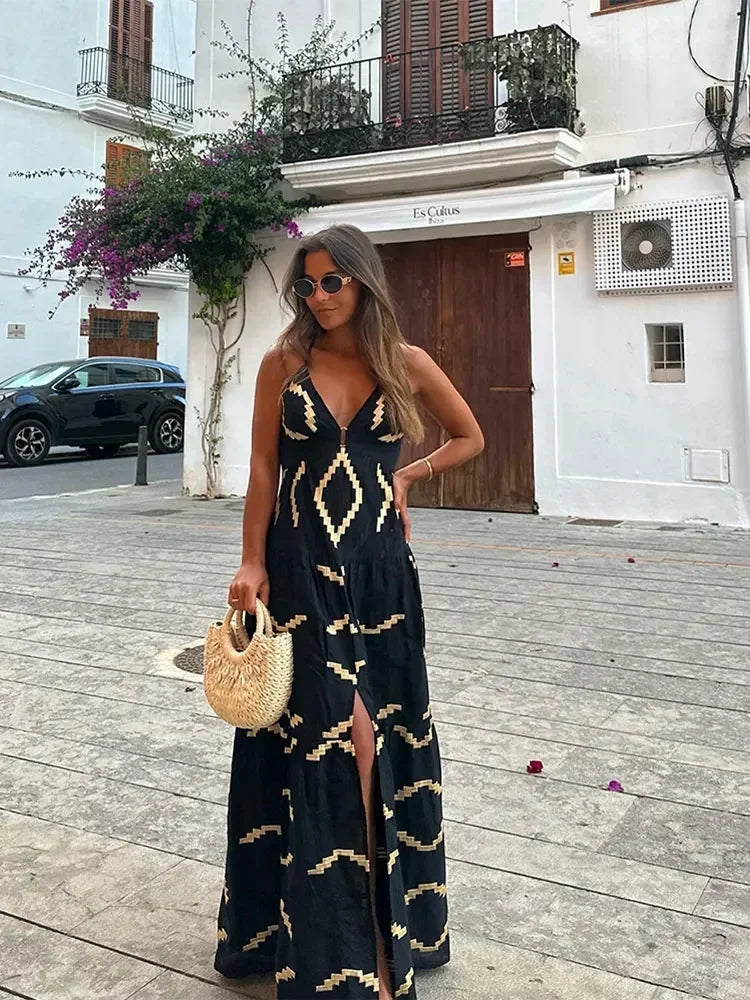 Woman in a black maxi dress with a gold geometric print, holding a straw bag and standing on a street with a building in the background.