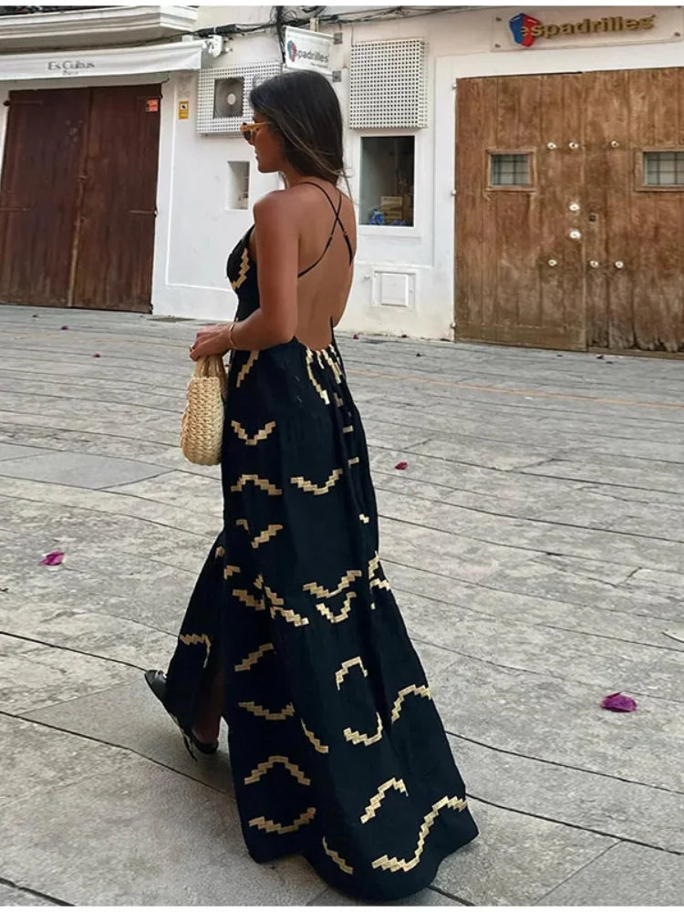 Woman in a black maxi dress with a gold geometric print, walking away from the camera on a street.