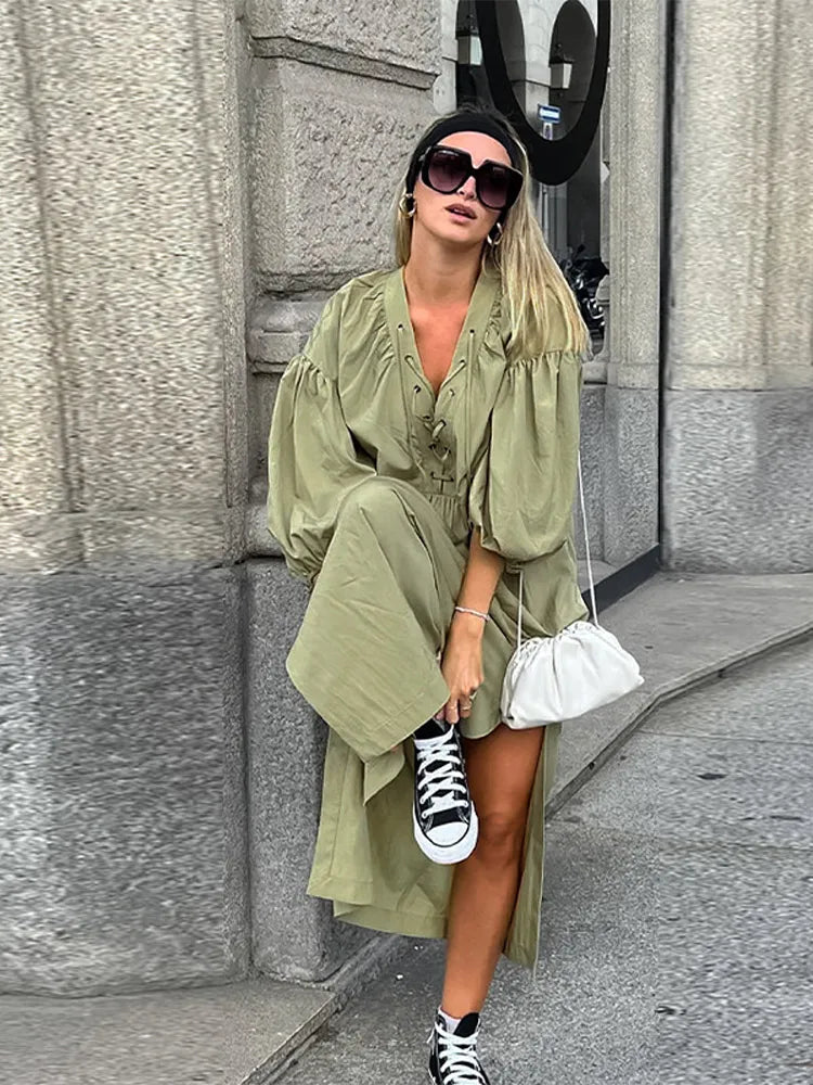 Woman in an olive green maxi dress with puff sleeves and a lace-up neckline, playfully lifting her leg to show off black high-top sneakers, carrying a white bag.