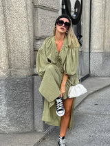 Woman in an olive green maxi dress with puff sleeves and a lace-up neckline, playfully lifting her leg to show off black high-top sneakers, carrying a white bag.