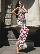A woman models a white maxi dress with a red rose print, posing with her hand on her face.