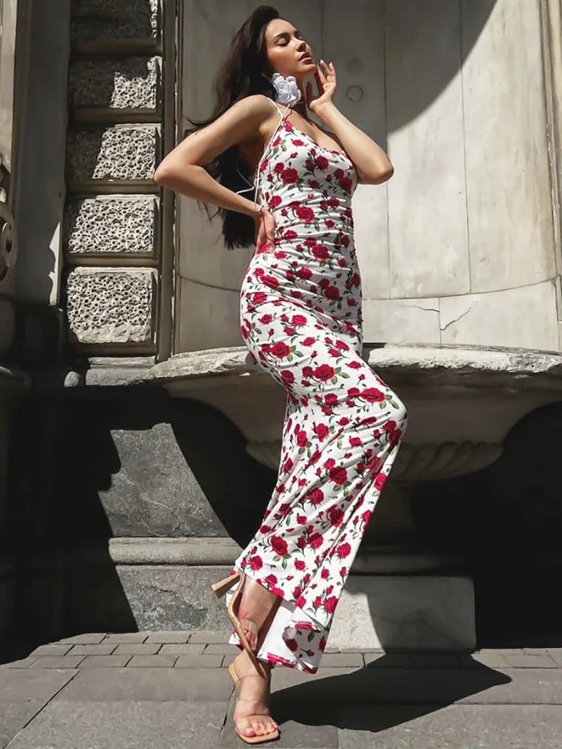 A woman models a white maxi dress with a red rose print, posing with her hand on her face.