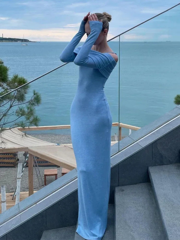 A young woman wearing a long, light blue off-the-shoulder dress with long sleeves, posing with her hands near her head, with the ocean in the background.