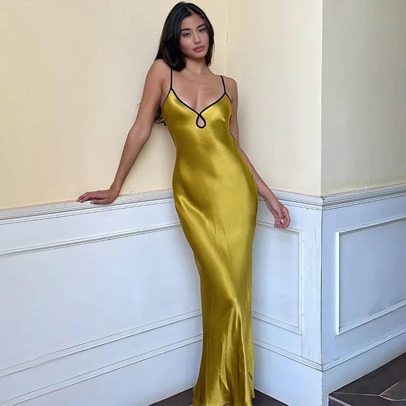 A woman leans against a wall wearing a long, yellow, sleeveless dress with thin black straps and a black trim around the neckline. She is looking towards the camera.
