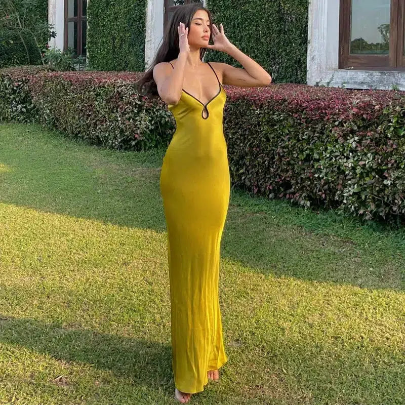 A woman stands in a garden wearing a long, yellow, sleeveless dress with thin black straps and a black trim around the neckline. She has her hands near her face.