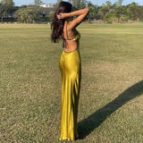 A woman stands in a grassy field wearing a long, yellow, sleeveless dress with thin black straps. The dress has an open back and is cinched at the waist.