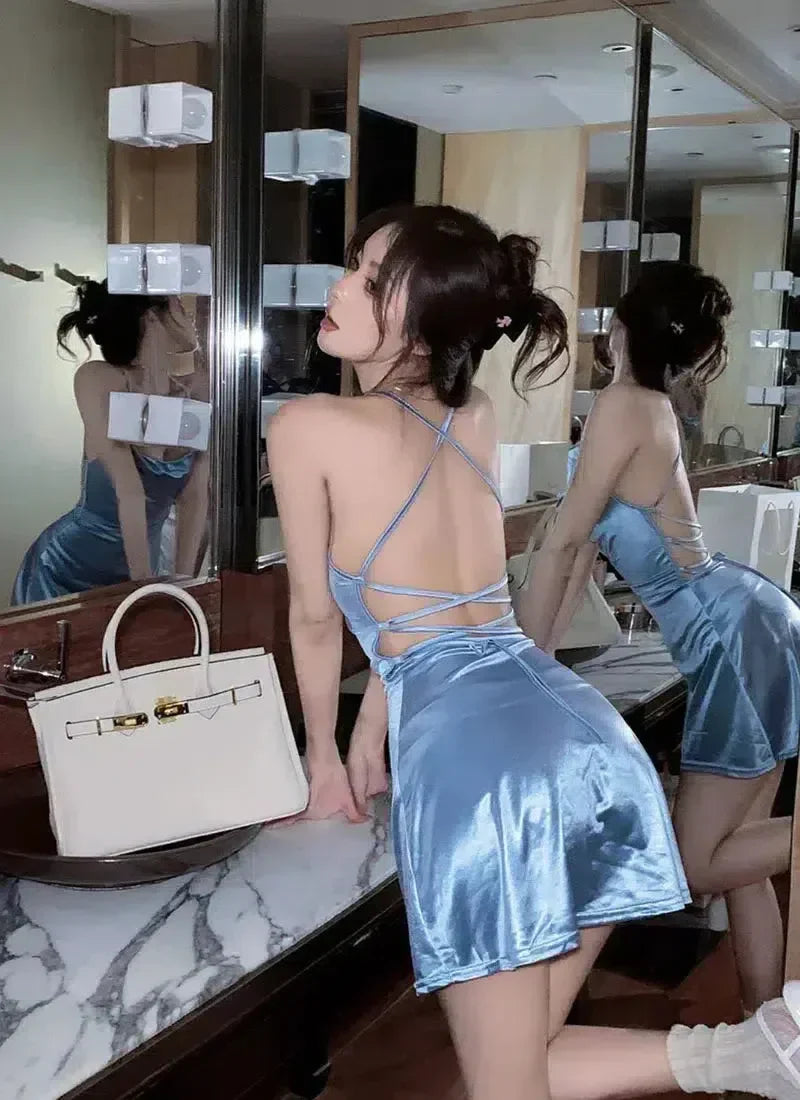 A woman in a light blue satin mini dress with a strappy back, posing in a bathroom.