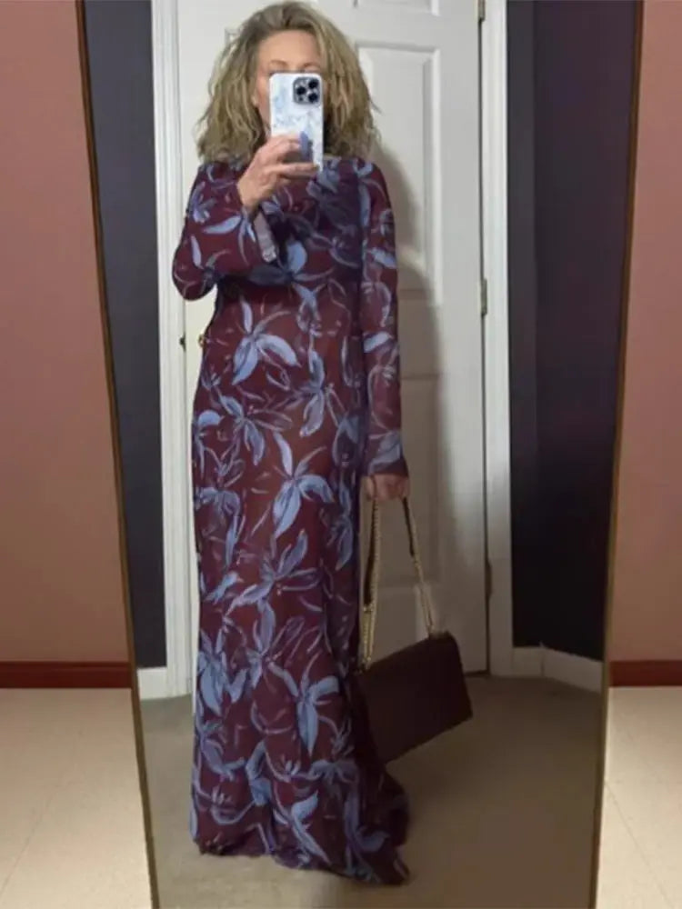 Woman in a sheer burgundy and blue floral print maxi dress, holding a handbag and taking a mirror selfie.