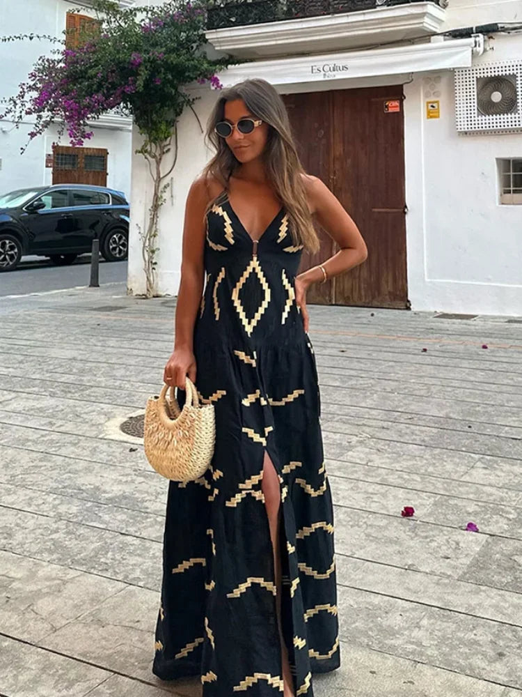 Woman in a black maxi dress with a gold geometric print, holding a straw bag and standing on a street.