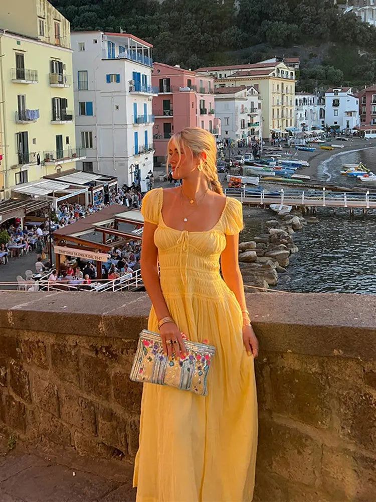 Woman in a yellow dress standing on a wall with a town and ocean in the background