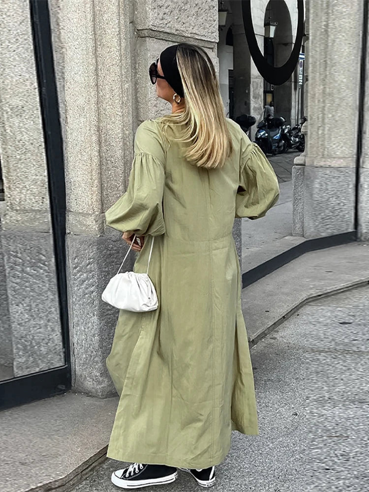 Woman in an olive green maxi dress with puff sleeves, showcasing the back of the dress with side slits, accessorized with a white bag and black high-top sneakers.