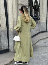 Woman in an olive green maxi dress with puff sleeves, showcasing the back of the dress with side slits, accessorized with a white bag and black high-top sneakers.