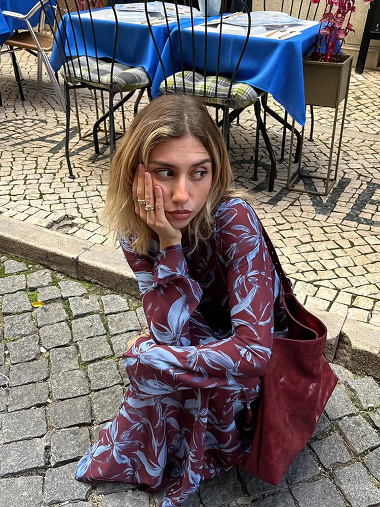 Woman wearing a long-sleeved burgundy and blue floral print maxi dress, sitting on cobblestones.