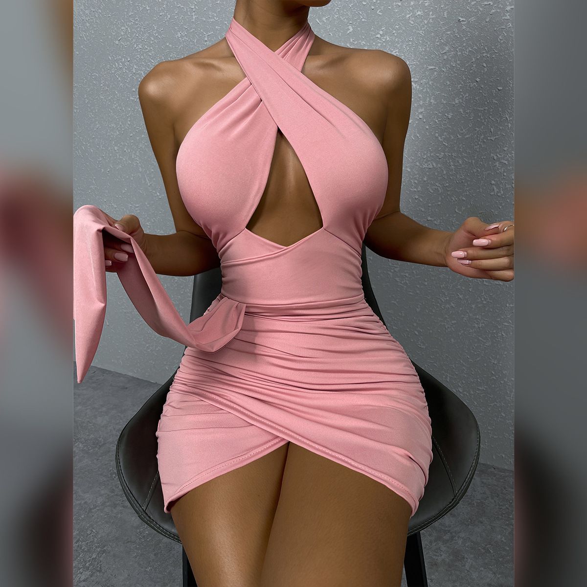 A woman in a pink halter neck dress with a cut-out and ruching is sitting on a chair.