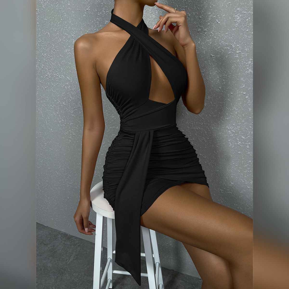 A woman in a black halter neck dress with a cut-out and ruching is sitting on a white stool.