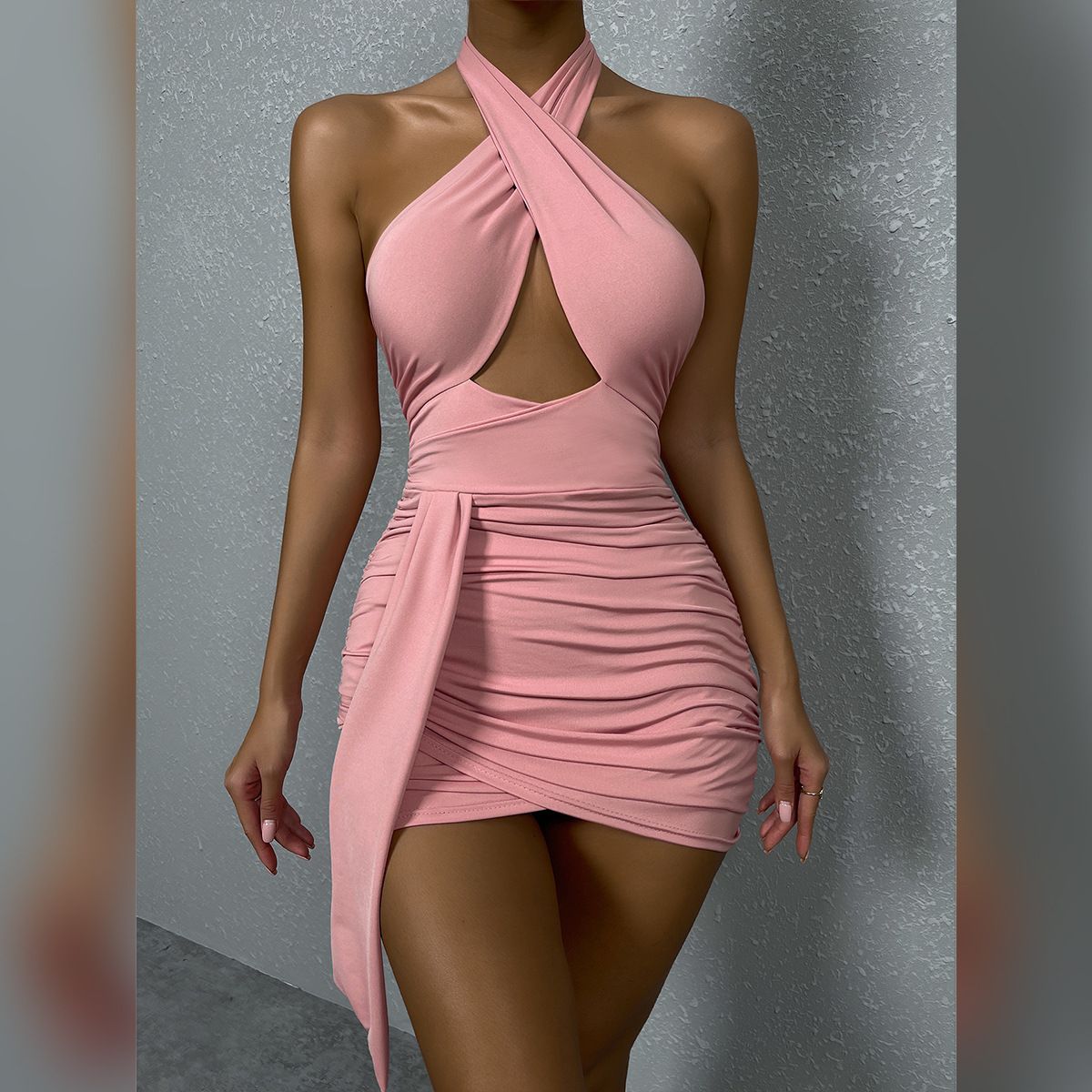 A woman in a pink halter neck dress with a cut-out and ruching is standing against a grey wall.