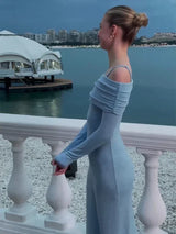 A young woman wearing a light blue off-the-shoulder dress with long sleeves, turned to the side, leaning on a white railing with a cityscape and ocean in the background.