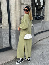 Rear view of a woman in an olive green maxi dress with puff sleeves, a lace-up neckline, and side slits, carrying a white bag and wearing black high-top sneakers.