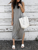 A woman in a grey sleeveless maxi dress with a knot detail at the hem and white slide sandals, standing against a weathered white wooden wall.