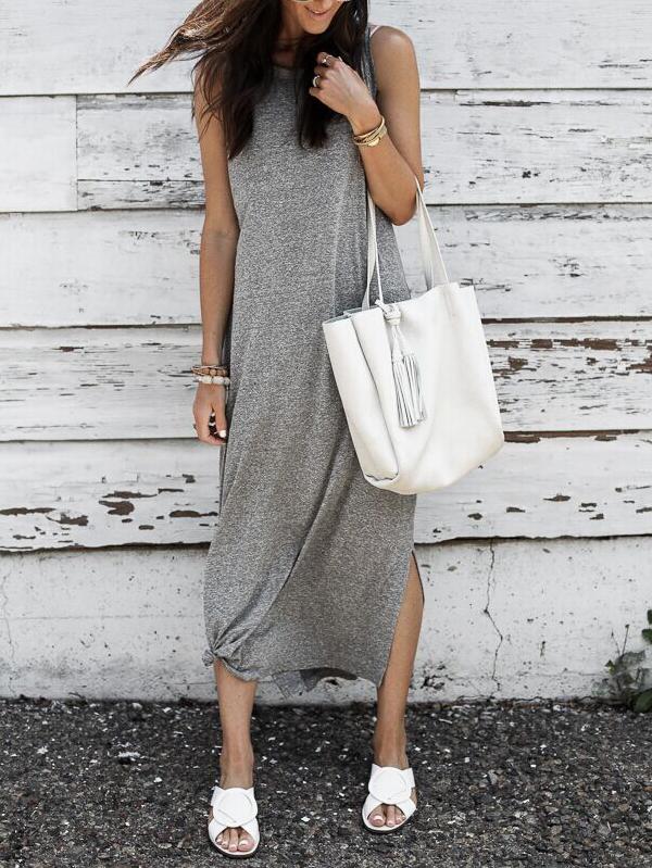 A woman in a grey sleeveless maxi dress with a knot detail at the hem and white slide sandals, standing against a weathered white wooden wall.