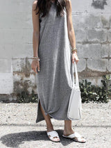 A woman in a grey sleeveless maxi dress with a knot detail at the hem and white slide sandals, standing in front of a brick wall.