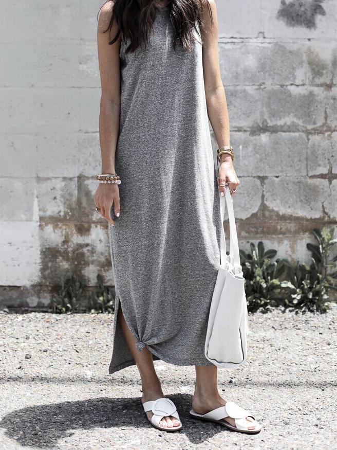 A woman in a grey sleeveless maxi dress with a knot detail at the hem and white slide sandals, standing in front of a brick wall.