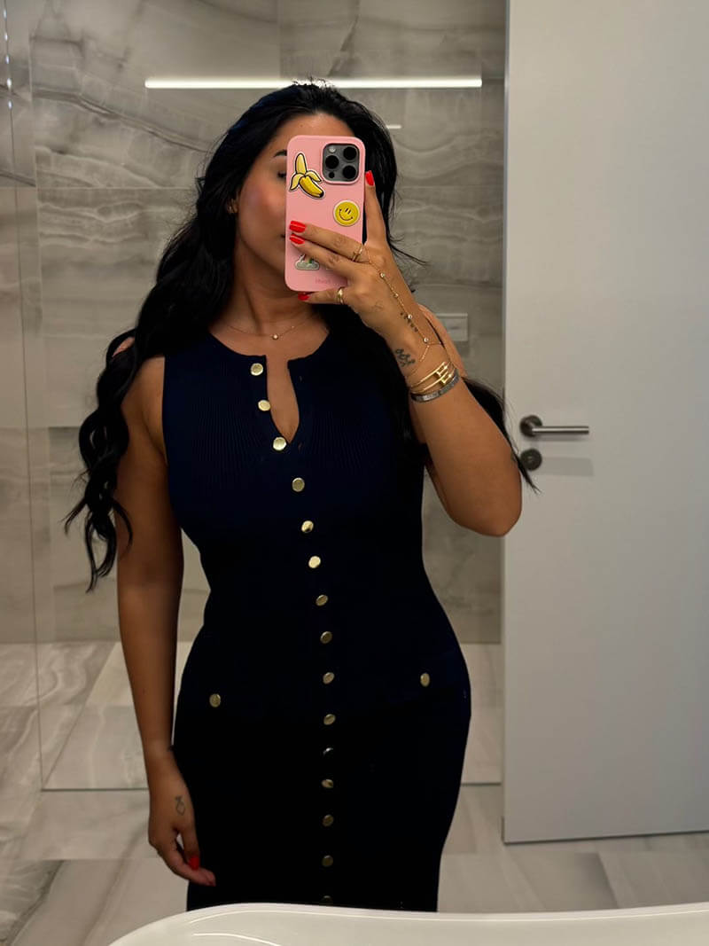 woman with long dark wavy hair wearing a navy ribbed knit sleeveless midi dress with gold buttons and pockets, standing in a bathroom