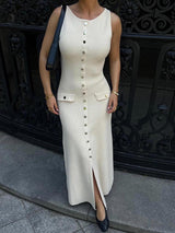 woman wearing a cream ribbed knit midi dress with gold buttons and pockets, black handbag, and black heels, standing in front of a black ornate gate