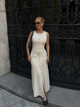 woman wearing a cream ribbed knit midi dress with gold buttons and pockets, black handbag, sunglasses, and black heels, standing in front of a black ornate gate
