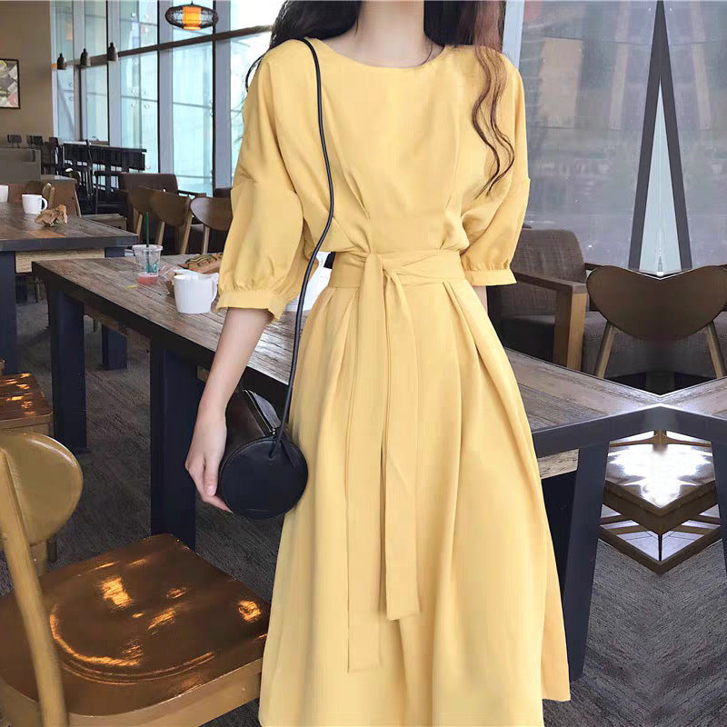 A woman stands in a cafe wearing a yellow midi dress with a tie waist and puff sleeves.