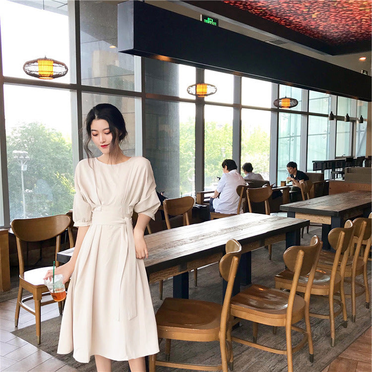 A woman stands in a cafe holding a drink, wearing a beige midi dress with a tie waist and puff sleeves.
