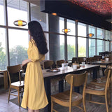 A woman stands looking out a window in a cafe, wearing a yellow midi dress with a tie waist and puff sleeves.