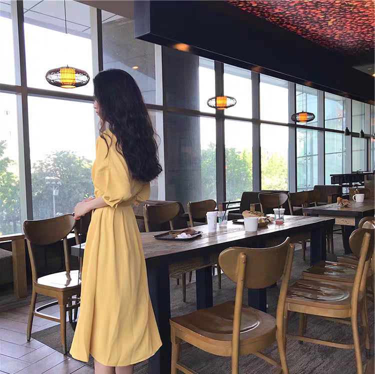 A woman stands looking out a window in a cafe, wearing a yellow midi dress with a tie waist and puff sleeves.
