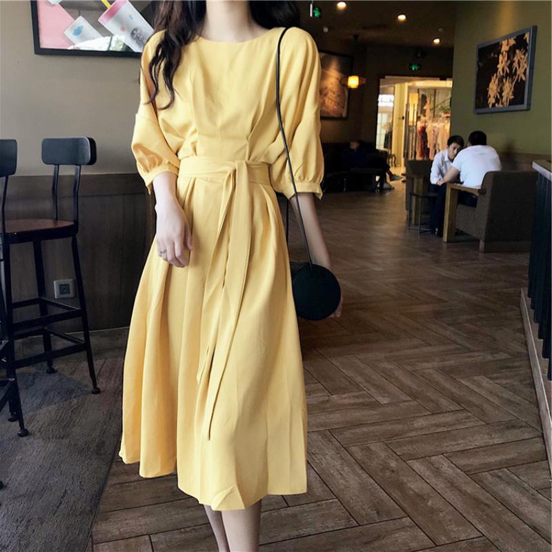 A woman stands in a cafe wearing a yellow midi dress with a tie waist and puff sleeves, with a black crossbody bag.