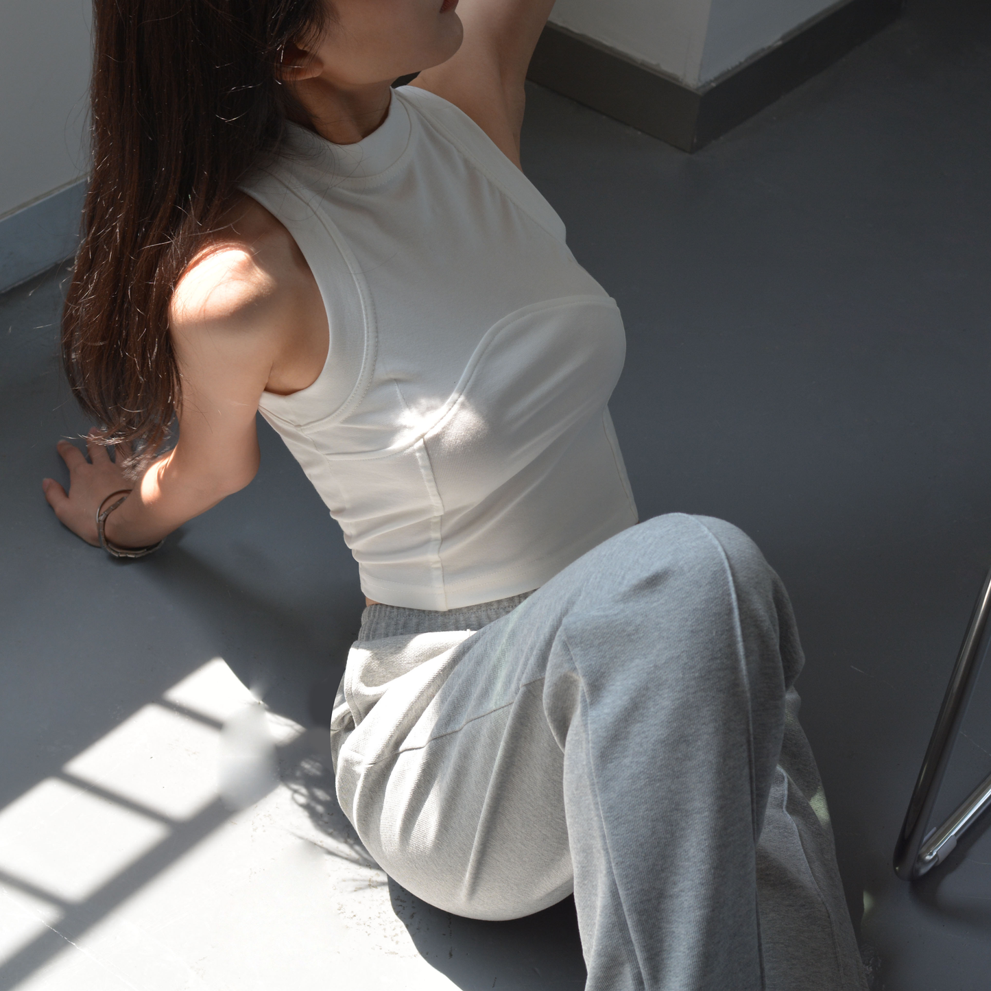 Woman wearing a white cropped tank top and grey sweatpants, sitting on the floor in sunlight.